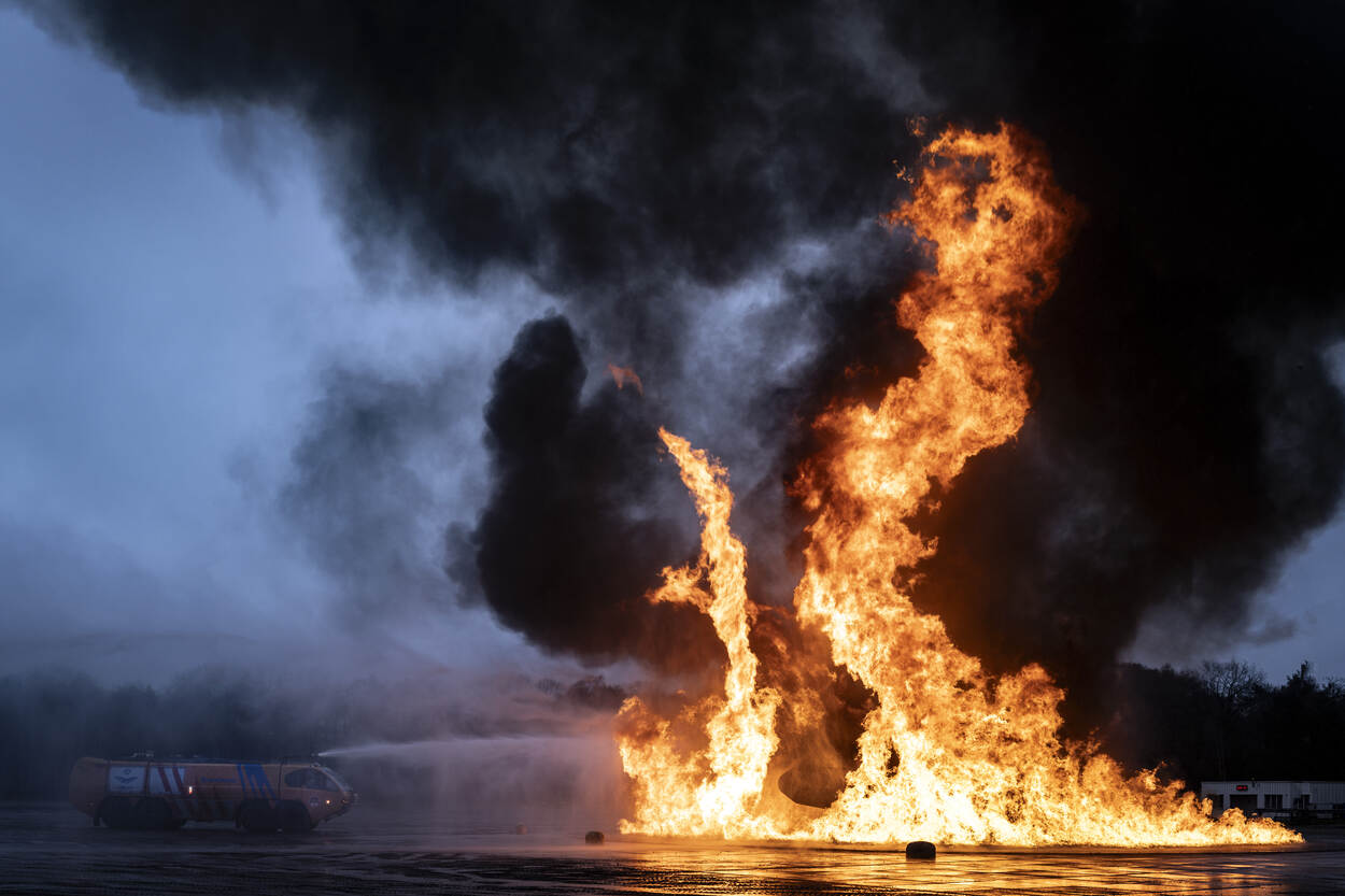 Enorme felgele en oranje vlammen schieten uit de mock-up van een verkeersvliegtuig met op de achtergrond gitzwarte rookwolken.