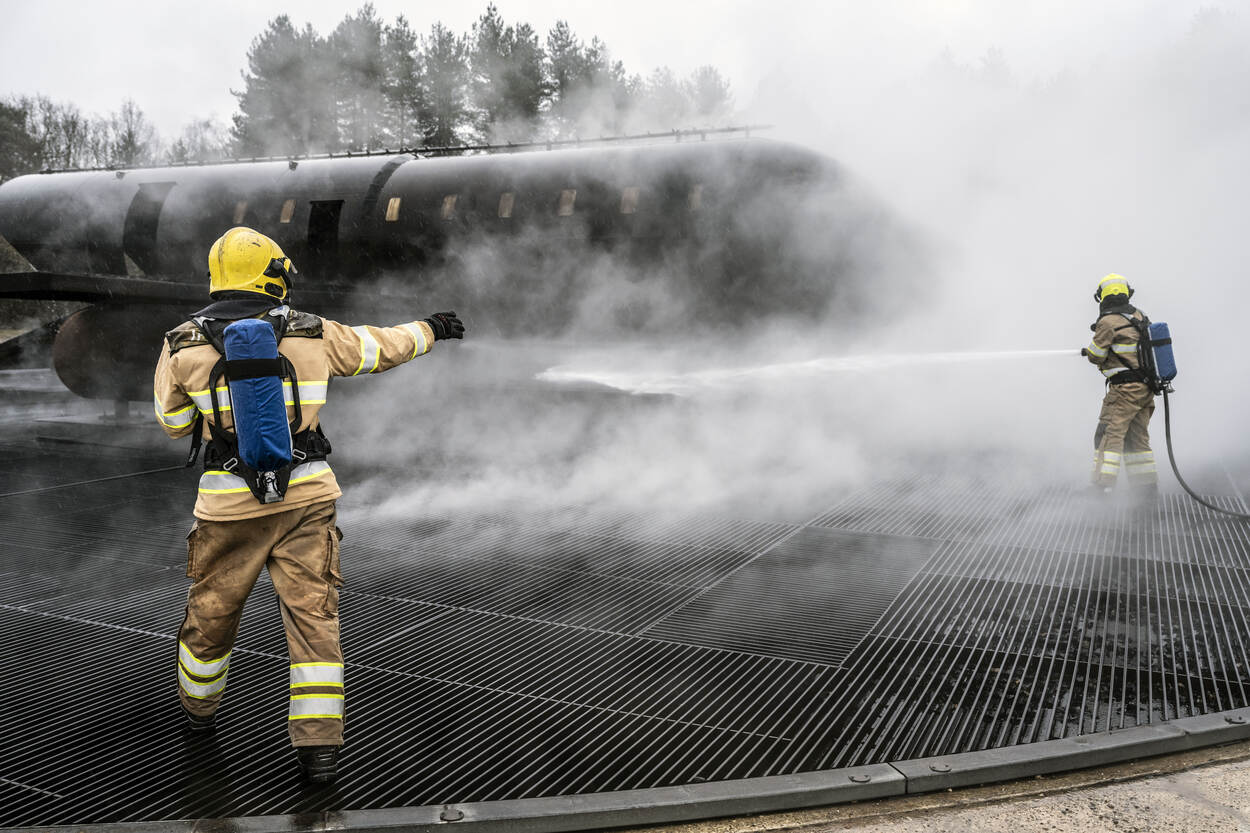 Twee brandweerlieden spuiten omringt door witte rook met een brandslang water op de mock-up.
