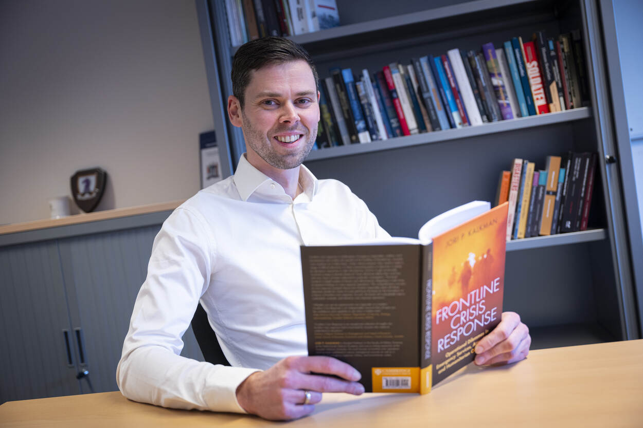 Auteur Jori Kalkman aan een tafel met in zijn handen zijn rood/oranje gekafte boek Frontline Crisis Response