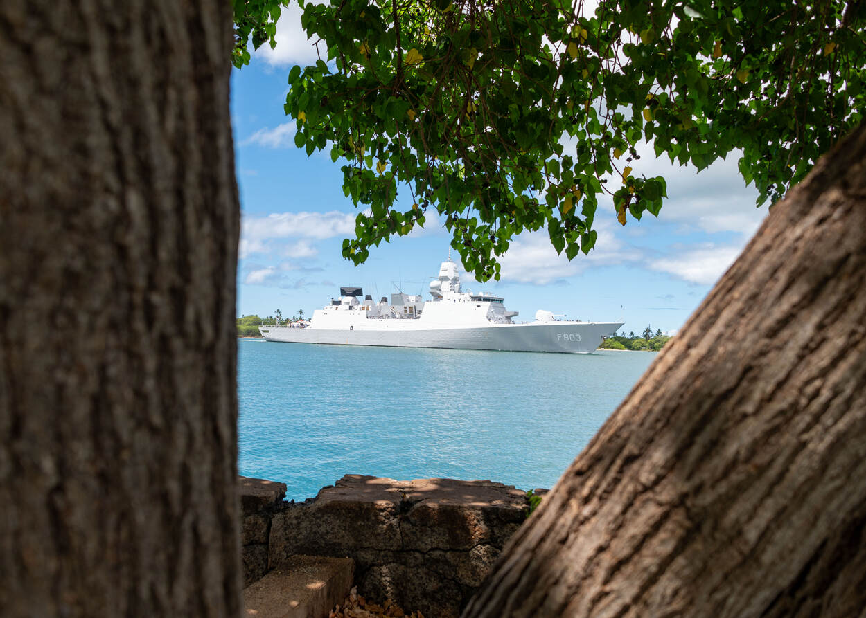 Tussen de bomen door zie je in de verte een Nederlands marineschip in een helderblauwe zee.