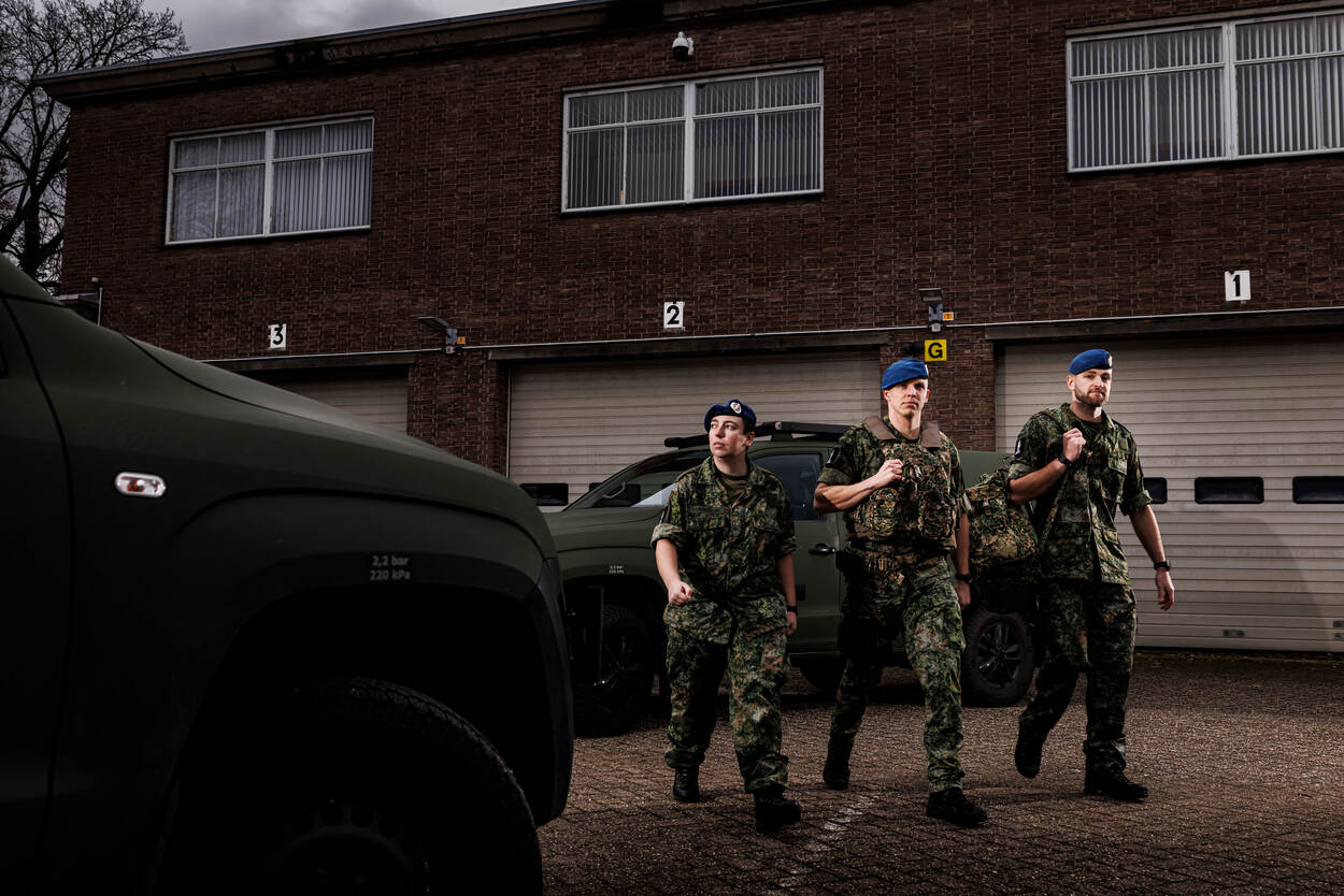 Drie militairen van de marechaussee, twee mannen en een vrouw, lopen in een groen camouflagepak langs voertuigen. Op hun hoofd de blauwe baret van de marechaussee.
