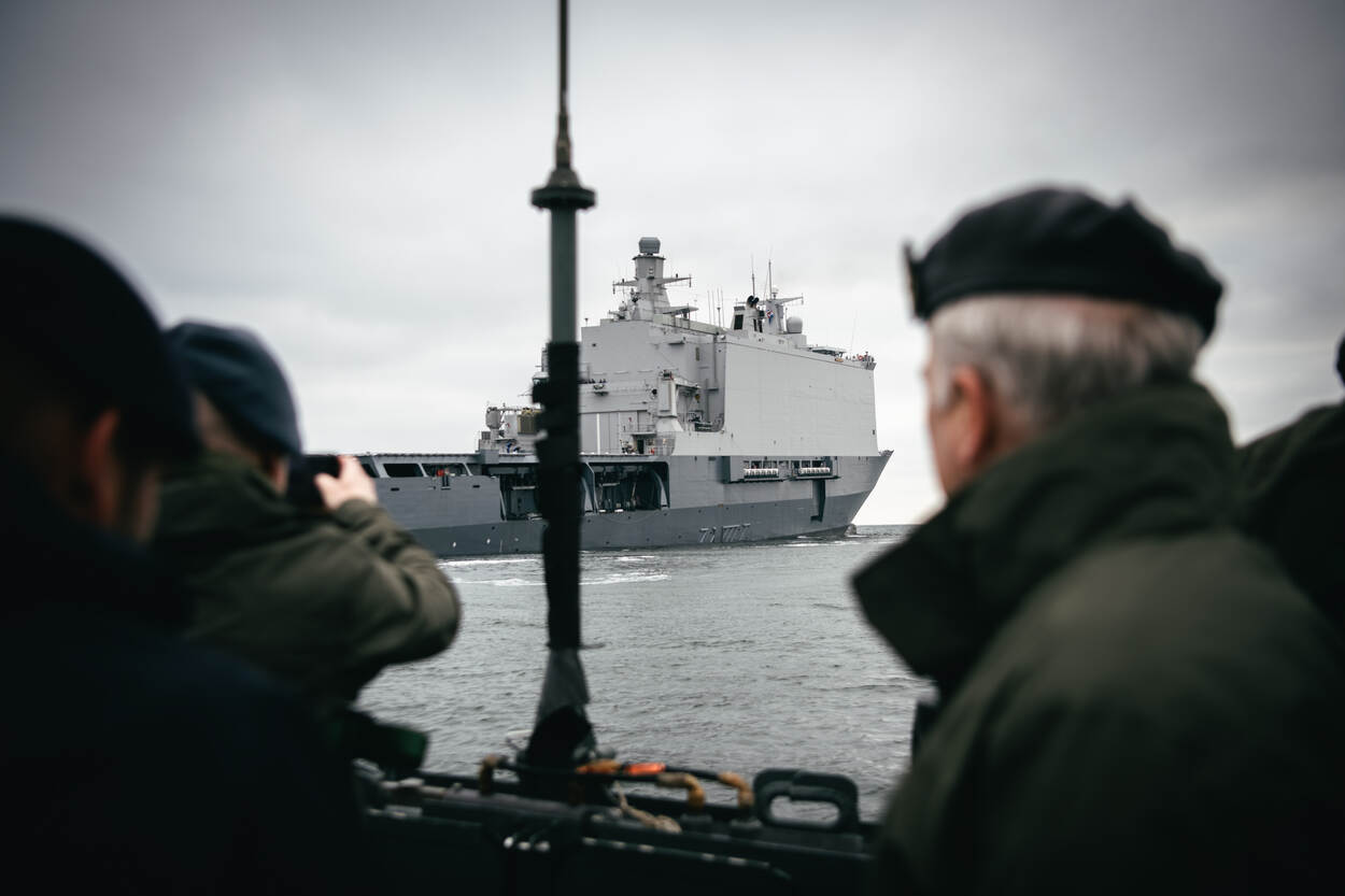 In de verte is amfibisch transportschip Zr.Ms. Johan de Witt op zee te zien. Op de voorgrond staan 2 militairen, van de achterkant bezien. Een van hen maakt een foto van het schip.