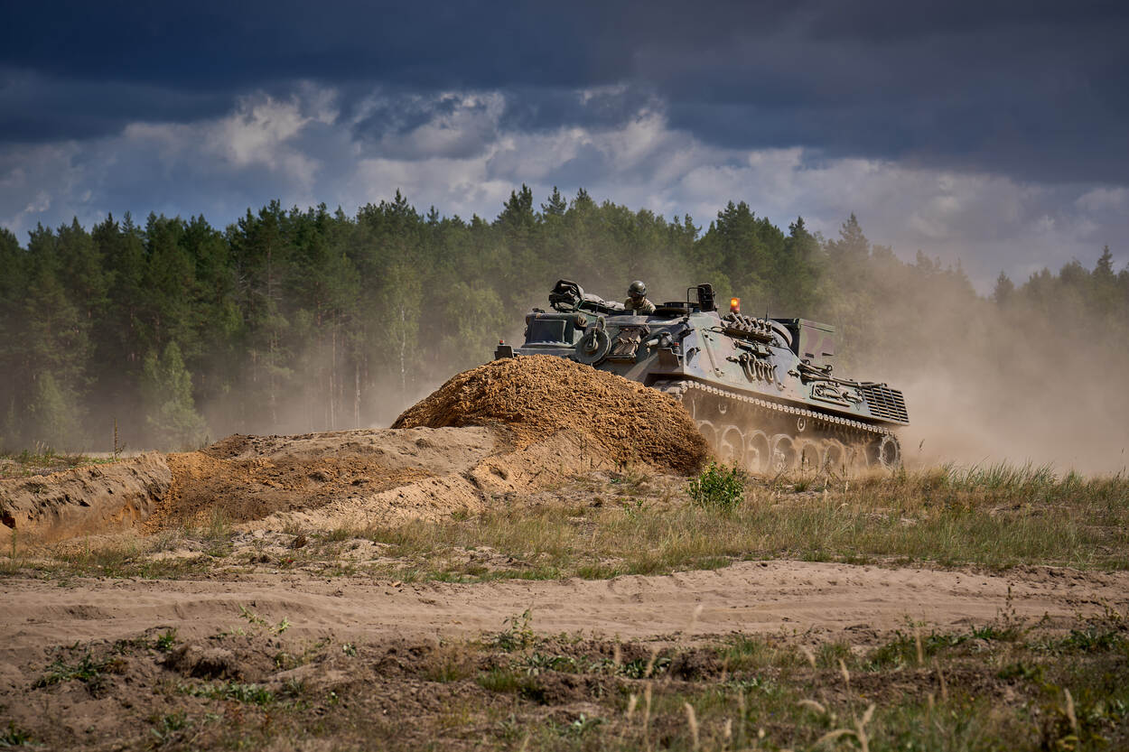 Een tank rijdt door een hoop zand. Op de achtergrond is de bosrand te zien.