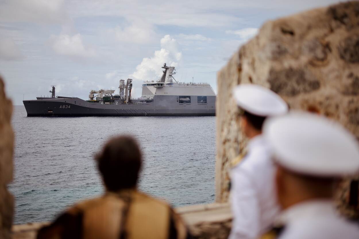 Twee marine-officieren in witte uniformen kijken naar het grote marineschip op zee, met een burger tussen rotsen op de voorgrond.