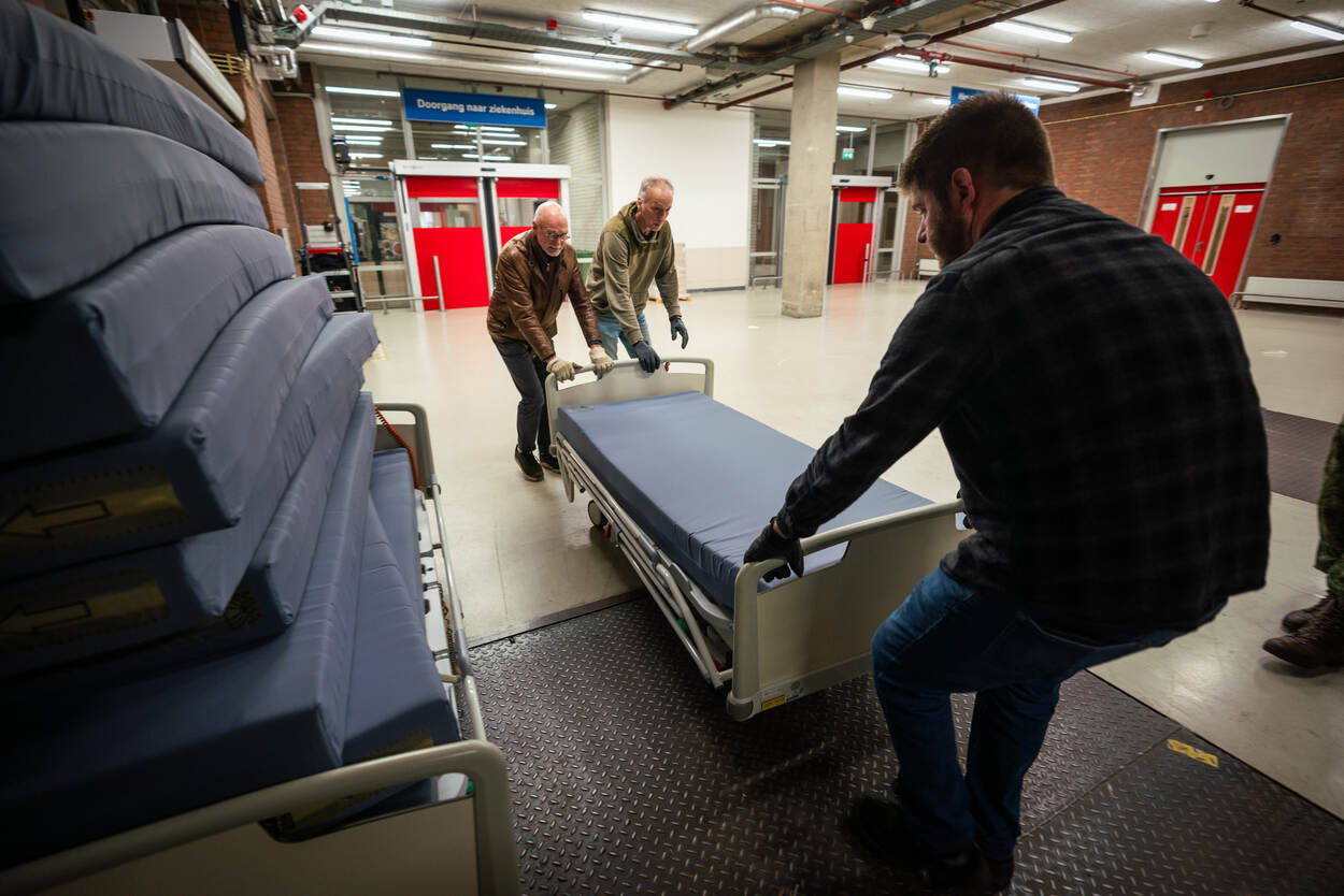 Drie mannen duwen en slepen een ziekenhuisbed in het laadruim van een vrachtwagen.