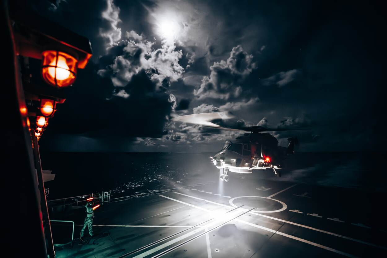 De NH90-boordhelikopter landt bij duisternis op het dek, waarbij er licht valt op de wolken.