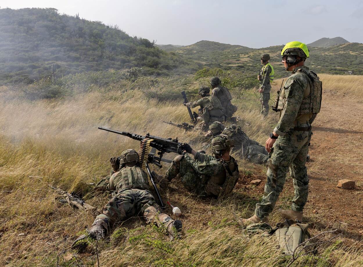Op de voorgrond ligt een groepswapenteam met een .50, dat doelen buiten beeld uitschakelt. Achter hen mortieristen met een 60 millimeter-mortier. Instructeurs kijken mee.