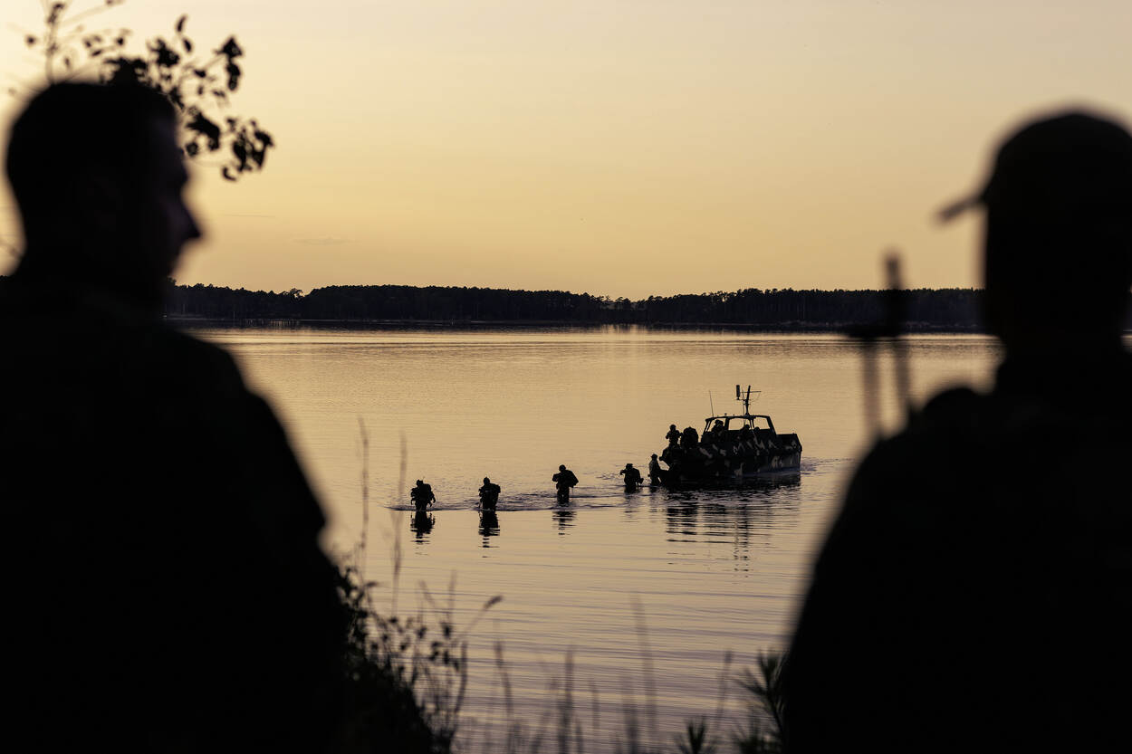 Mariniers lopen van de boot af, door het water, richting de wal, in de avondzon.