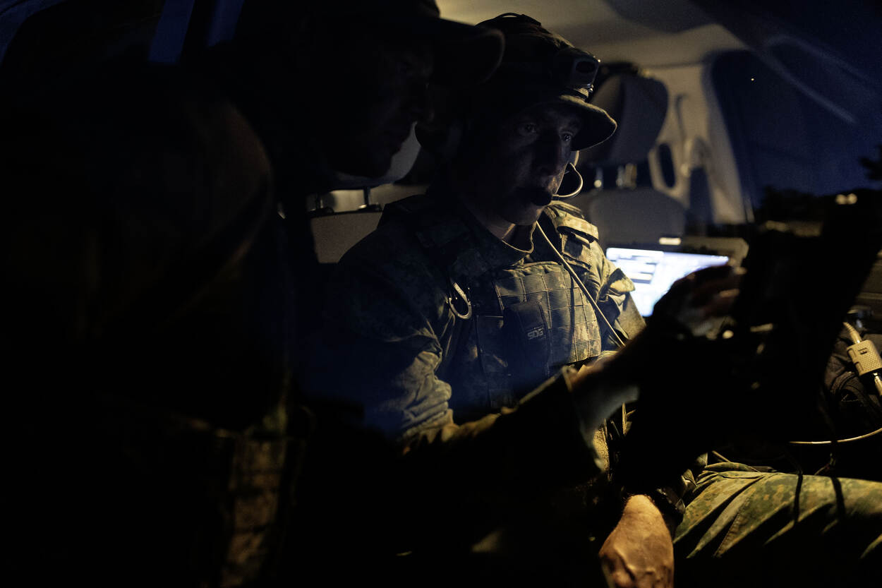 Een militair zit in een voertuig en kijkt in de duisternis naar een scherm. Zijn collega staat naast hem, buiten het voertuig, en kijkt mee.