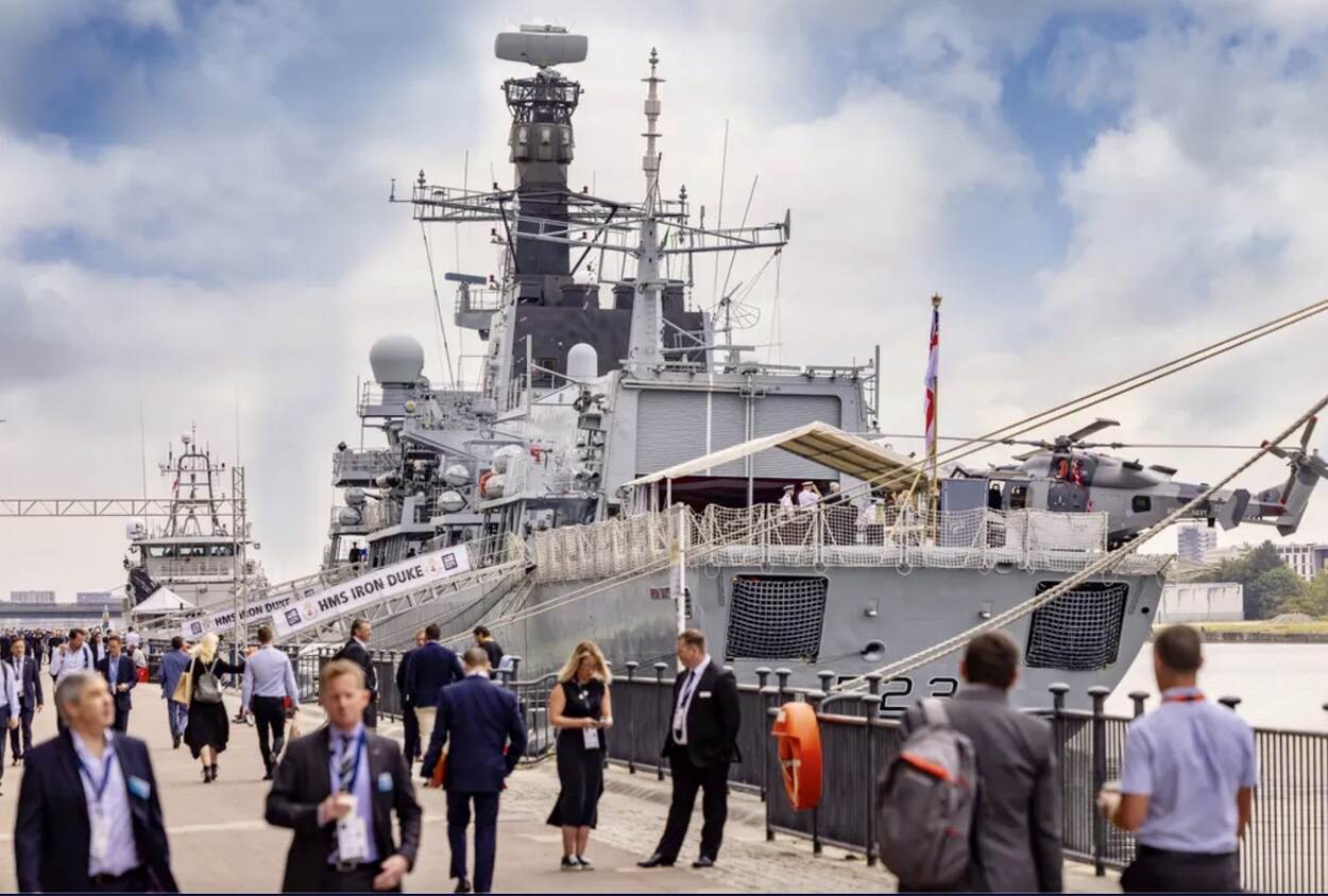 Een fregat aan de kade van London Excel, met een helikopter op het dek. Veel mensen kijken en lopen rond.