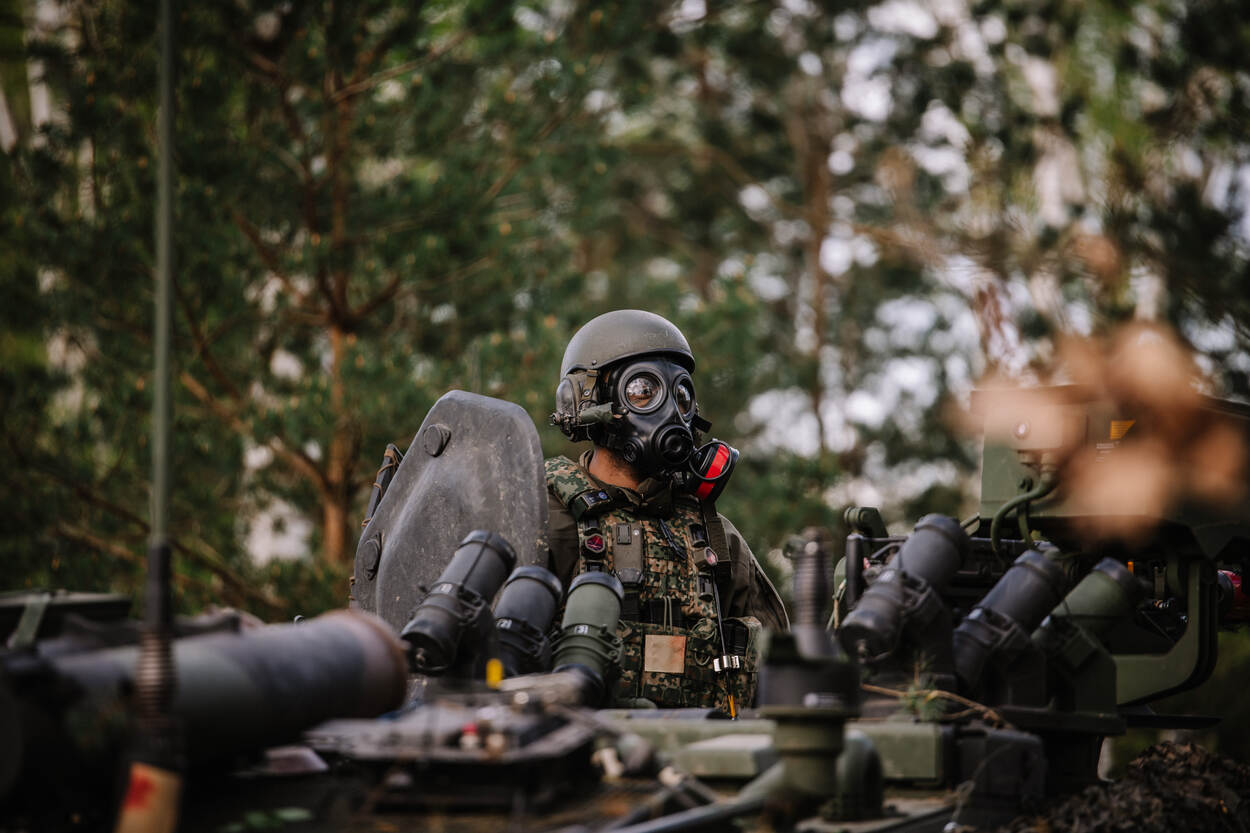 Een militair staat met een gasmasker op zijn hoofd in het veld. Voor hem staat een voertuig.