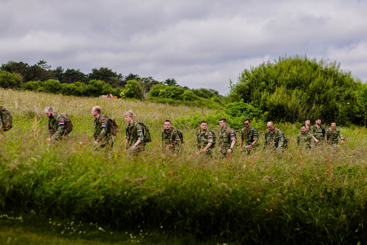 13 bepakte fuseliers lopen in een rij door het hoge gras.