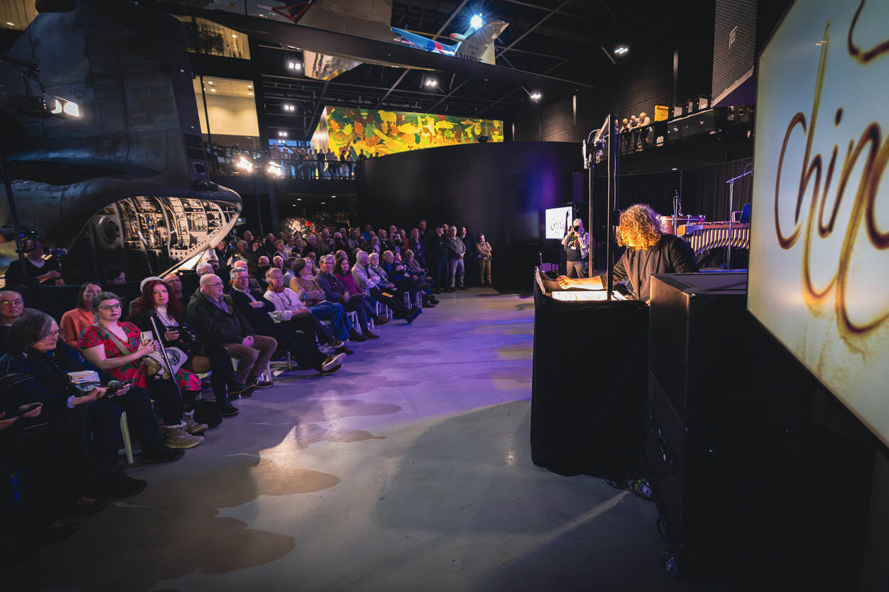 Overzicht van links publiek dat voor een Chinook zit, en rechts de zandtovenaar en een tv-scherm waarop groot te zien is wat hij met zand maakt.