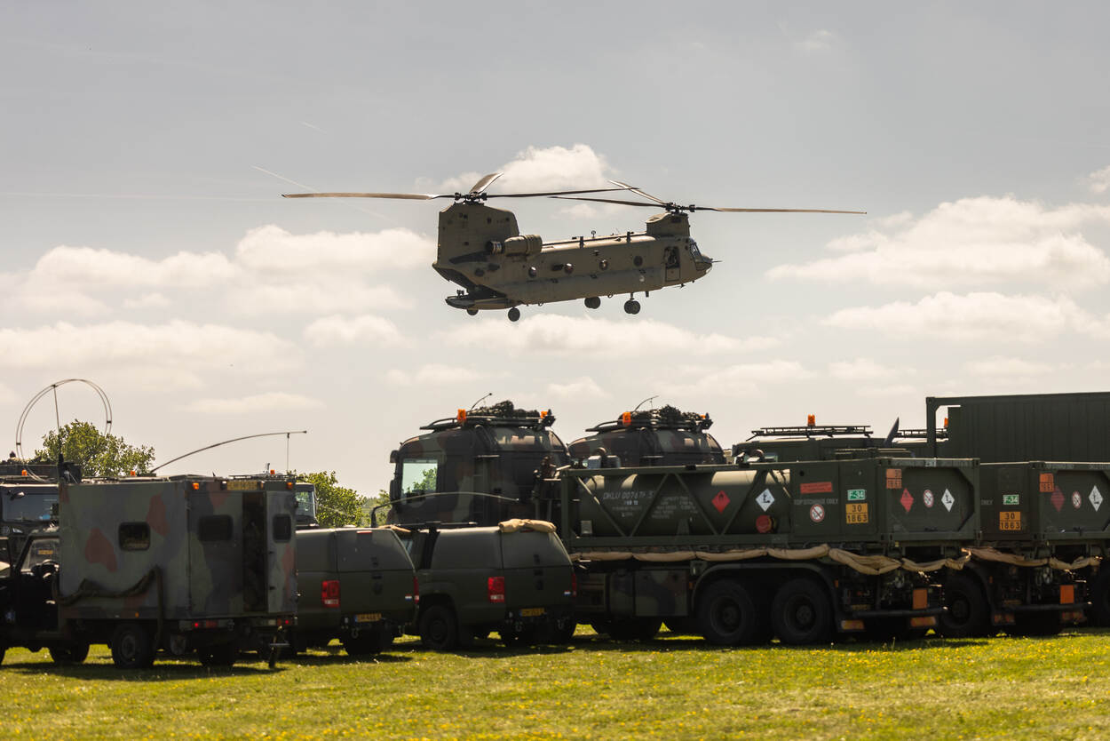 Chinook vliegt laag over geparkeerde militairen voertuigen op het gras.