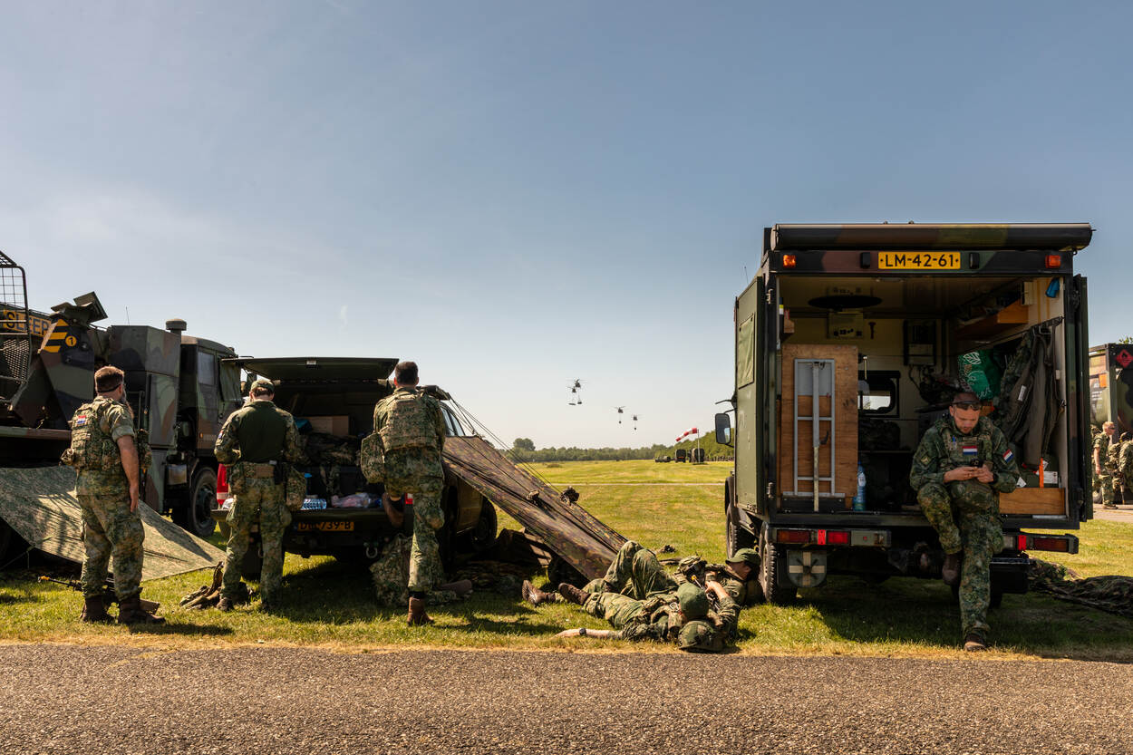 Voertuigen en militairen op een grasveld. Naast een voertuig een gespannen poncho als slaaponderkomen. In de verte helikopters met slingload.