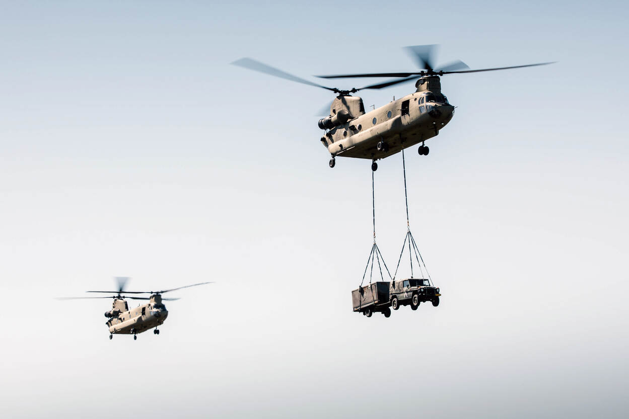 Twee Chinooks vliegen achter elkaar door de lucht. Een daarvan verplaatst een auto aan een zogenoemde sling onder de helikopter.