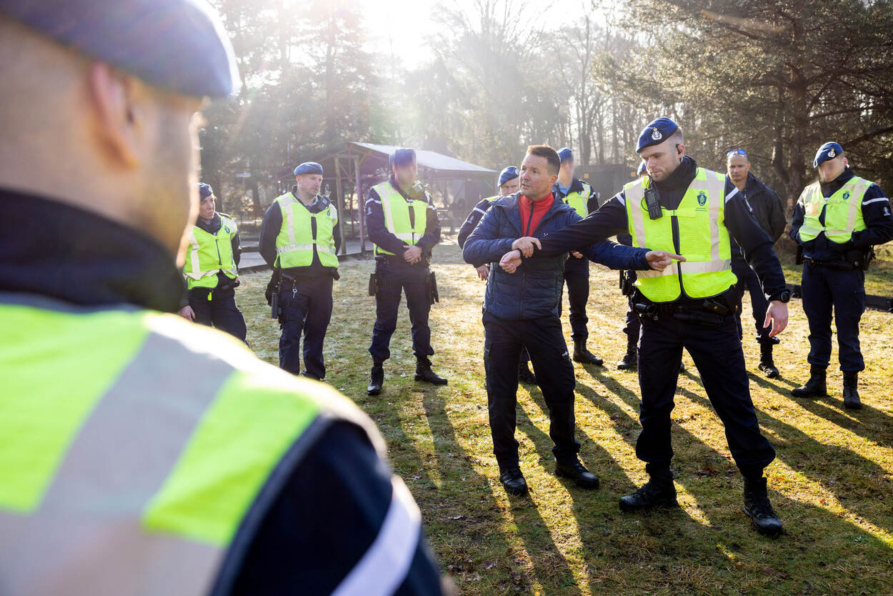 Een groep marechaussees met gele veiligheidshesjes staat in een cirkel rond een instructeur van de politie. De instructeur houdt de arm van een van de collega’s vast om een handeling voor te doen.