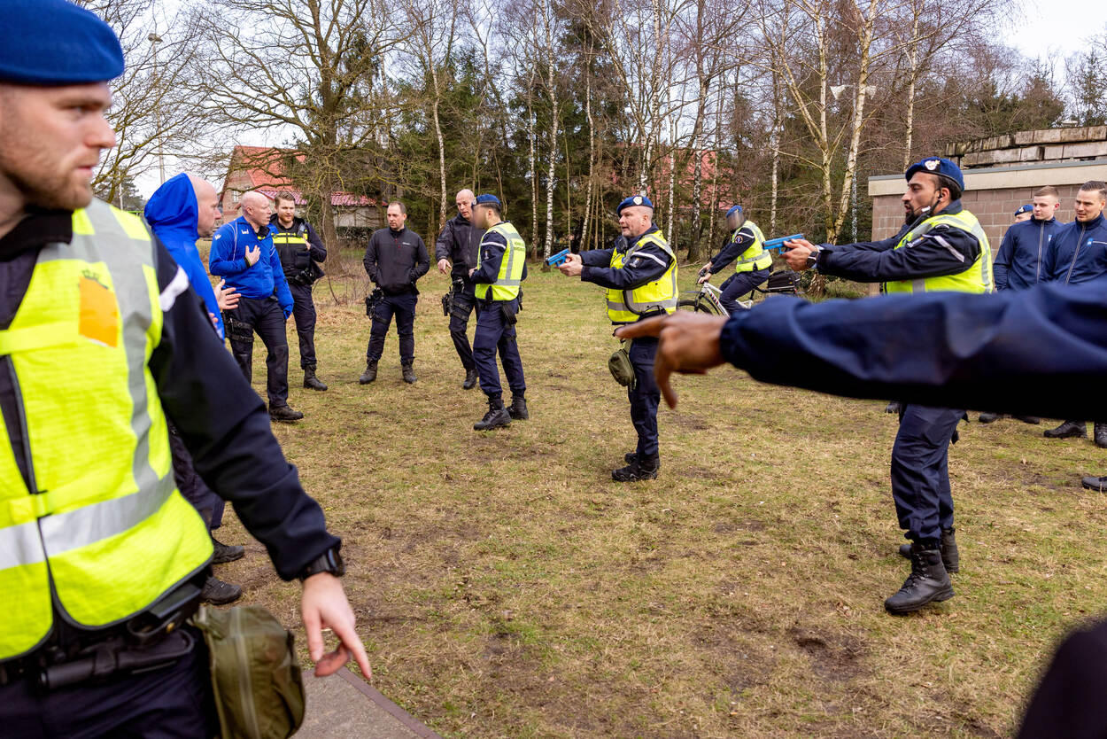 Drie marechaussees trekken hun oefenwapen, daaromheen staat een groep omstanders.