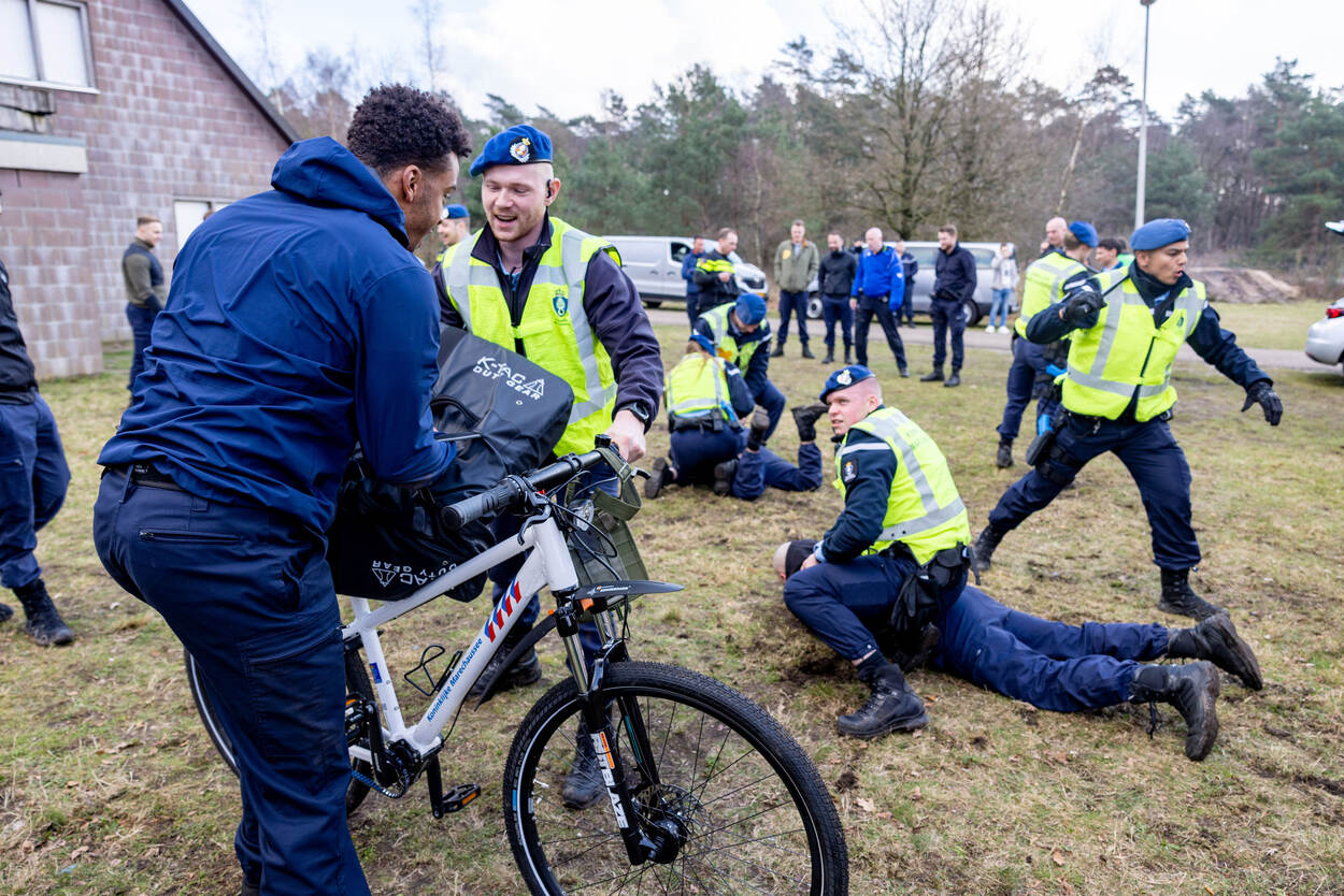 Marechaussees met geboeide tegenspelers. Een collega blokkeert de toegang voor omstanders met zijn witte fiets.