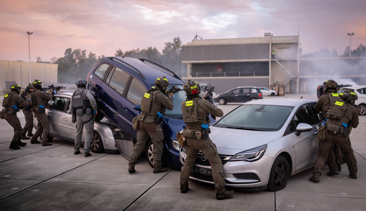 In uniform gehulde deelnemers van verschillende landen schieten met hun wapens van achter een aantal auto’s.