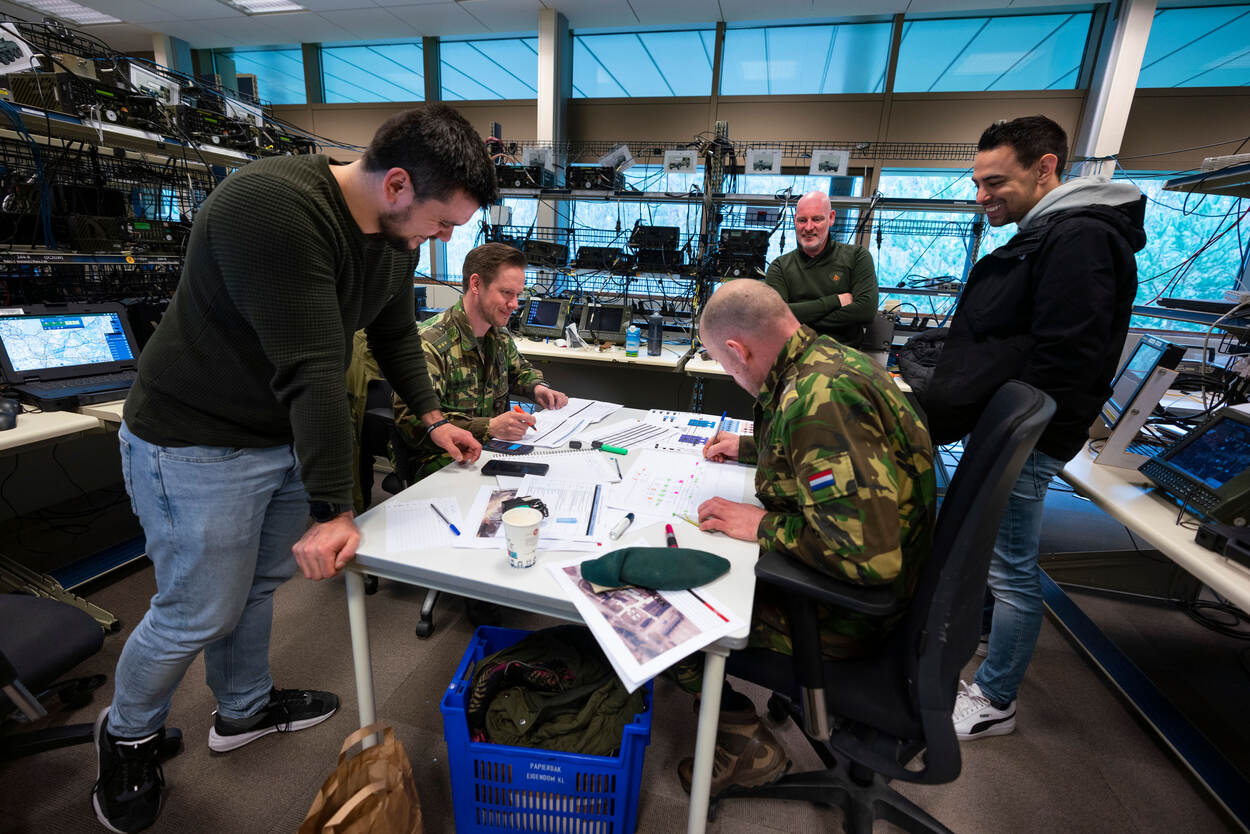 Burgermedewerkers van JIVC en militairen overleggen aan een tafel tussen kasten met radio's.
