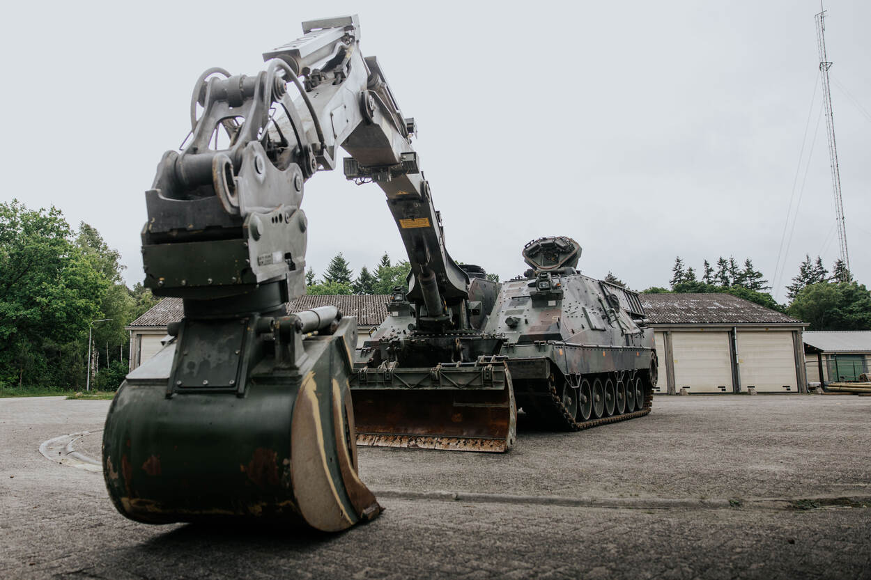 Een geparkeerde Kodiak. Gezien vanaf de voorzijde. De graafarm rust op de grond. Achter het geniedoorbraaksysteem zijn loodsen te zien.