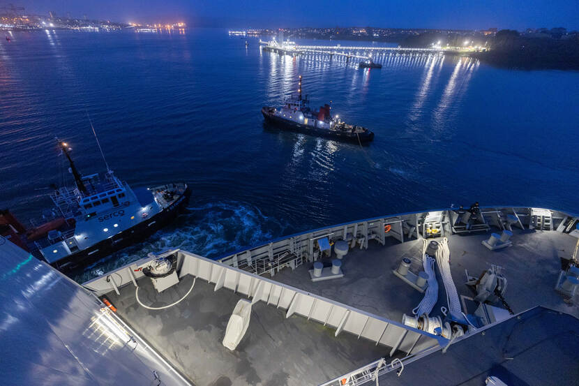 De Karel Doorman wordt begeleid door twee sleepboten, met op de achtergrond een verlichte oliesteiger.