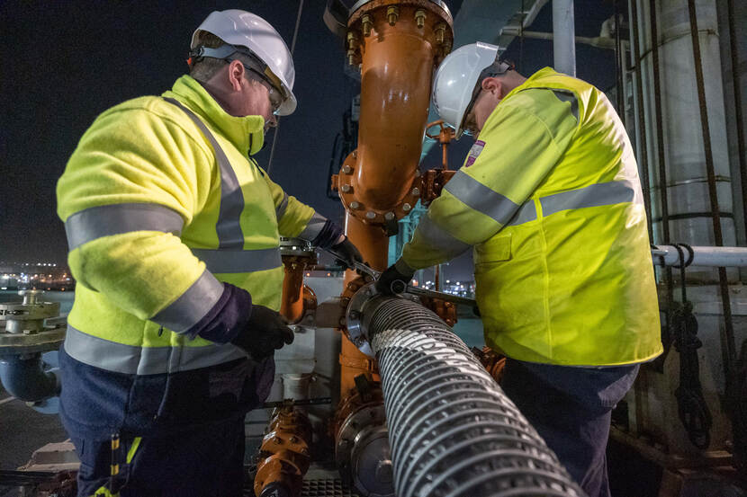 Twee personen in gele veiligheidsjassen draaien de moeren vast die de tankslang aan het schip koppelen.