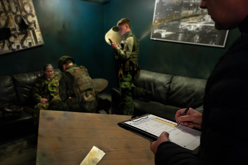 Op de andere afbeelding staan we in dezelfde ruimte, maar kijken we mee naar het beoordelingsformulier van een van de instructeurs, die de inzet een cijfer geeft.