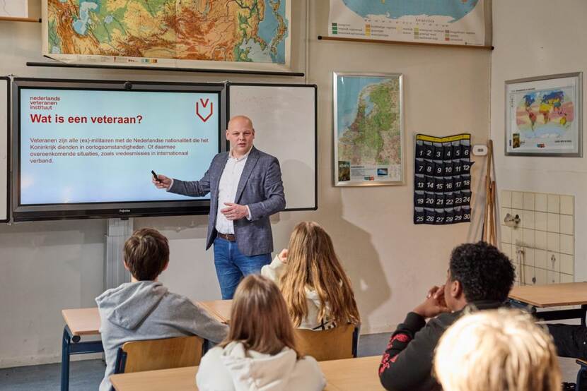 Man staat voor een klas en spreekt kinderen toe terwijl hij naar informatie op het schoolbord wijst