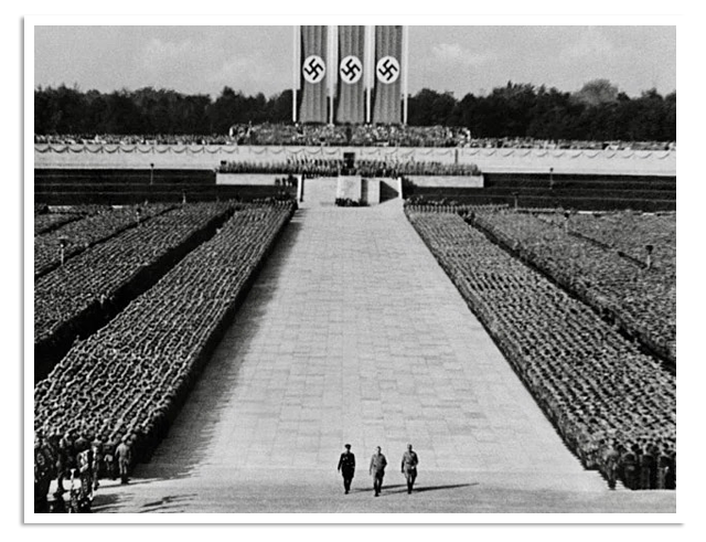 Rijen met soldaten staan opgesteld in een arena, terwijl drie man naar een podium lopen voor een toespraak.