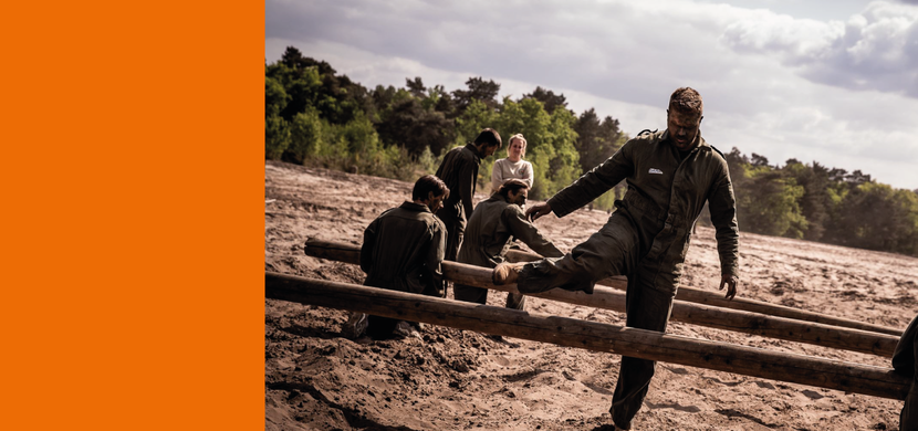 Een groep militairen in een groene overall rent door het zand en springt over balken.