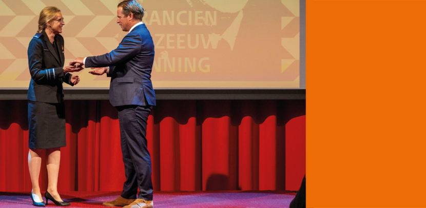 Generaal Boekholt-O’Sullivan krijgt de penning overhandigd door staatssecretaris Gijs Tuinman. Beiden dragen een blauw pak en glimlachen.