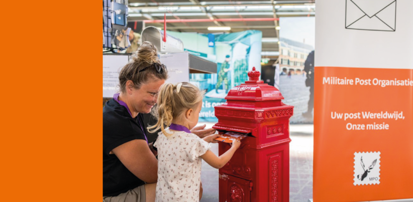 Een blonde dame met een knot op haar hoofd staat voor een rode brievenbus met een klein meisje. Samen stoppen ze een brief in de brievenbus. Daarnaast staat een banner met de tekst ‘Militaire Post Organisatie. Uw post, wereldwijd, onze missie.”