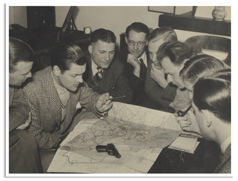 Zwart-witfoto van een groep jonge mannen. Op tafel liggen een landkaart en een revolver.