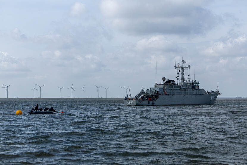 Vanuit een rubber bootje zetten duikers een boei uit, rechts is BNS Lobelia te zien en op de achtergrond de kust van Eemshaven met een aantal windturbines.