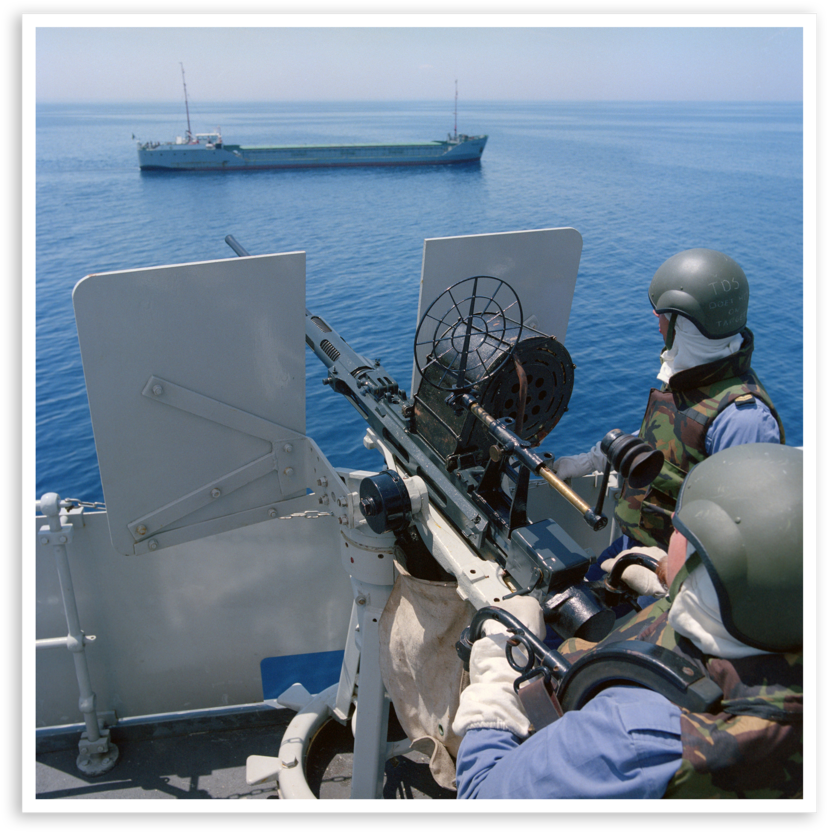 Twee militairen staan gereed achter een 20mm-Oerlikon-mitrailleur. Verderop vaart een schip, waarop het wapen gericht is.