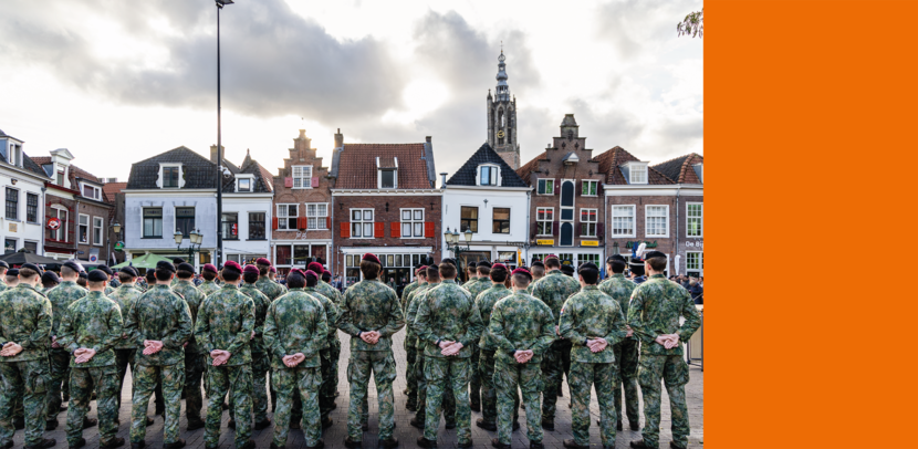 Een groep mensen in militaire kleding staat in gelid, met historische gebouwen en een kerktoren op de achtergrond onder bewolkte lucht.