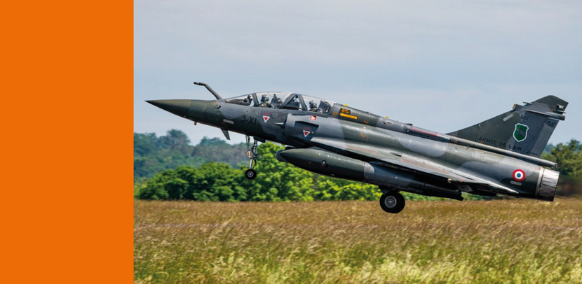 Een militair straaljagertoestel tijdens het opstijgen vanaf een grasveld, met inzetbare landingsgestel en rookontwikkeling achter de motoren.