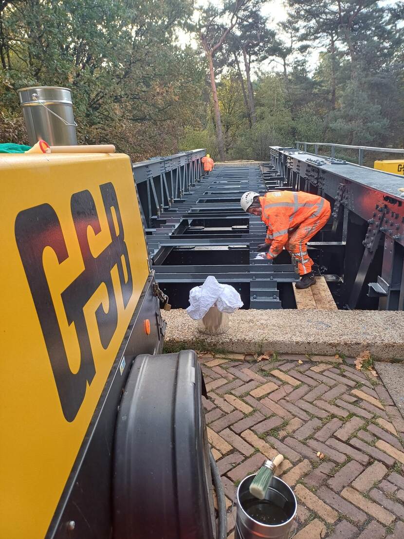Een bouwwerker in een oranje veiligheidspak staat op een metalen constructie, terwijl een gele machine met de letters GSB erop op de voorgrond staat.