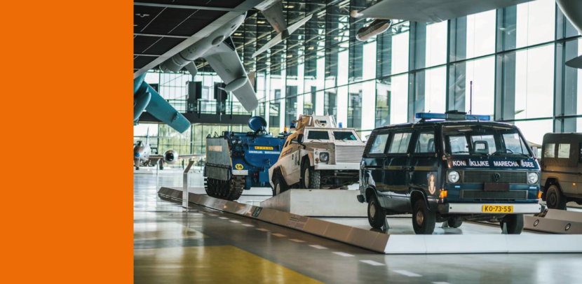 Historische voertuigen van de Koninklijke Marechaussee tentoongesteld in een museumhal, met pantserwagens en een dienstbus onder vliegtuigen.