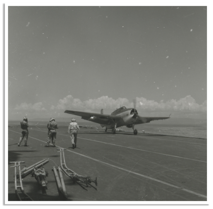 Een oud militair vliegtuig staat op het dek van een vliegdekschip, met drie bemanningsleden eromheen en een bewolkte lucht op de achtergrond.