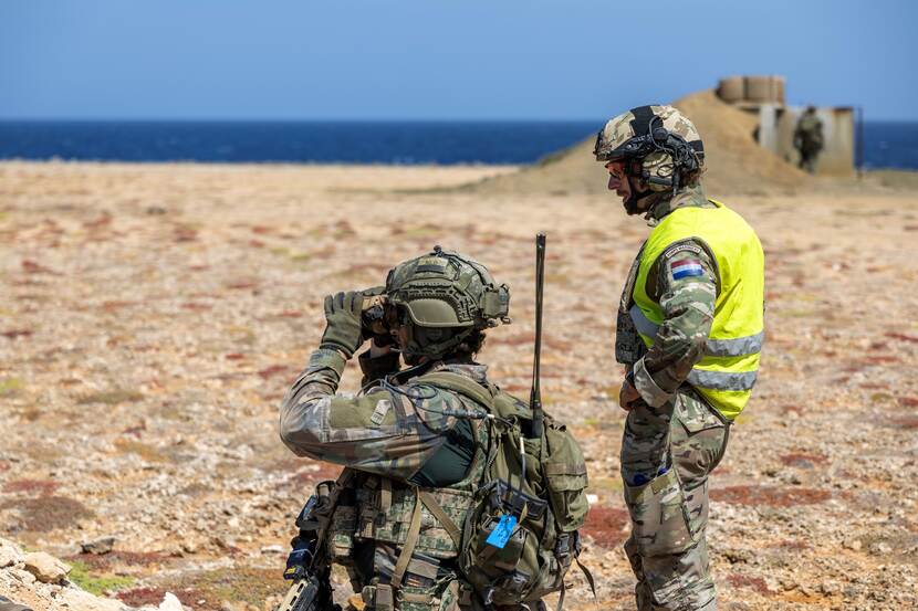 Een gevechtsschietinstructeur met een geel hesje. Naast hem een geknielde marinier die door een verrekijker kijkt.