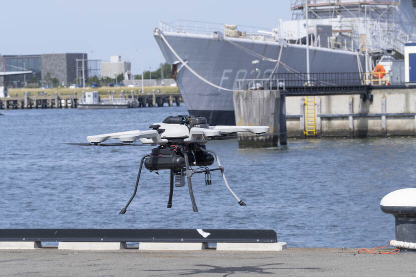 Een vliegende drone. Op de achtergrond ligt een marineschip aan de kade.