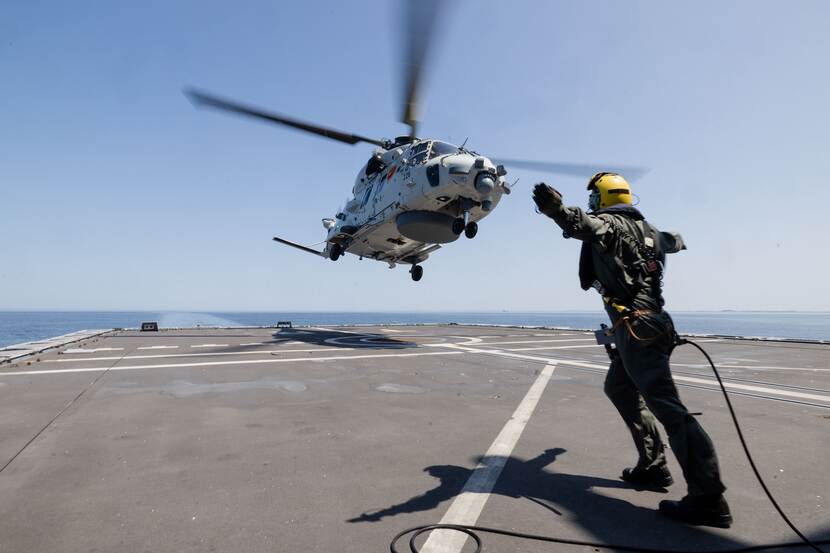 Een NH90-boordhelikopter stijgt op van het helidek van de Tromp onder begeleiding van de vliegdekofficier op de voorgrond.