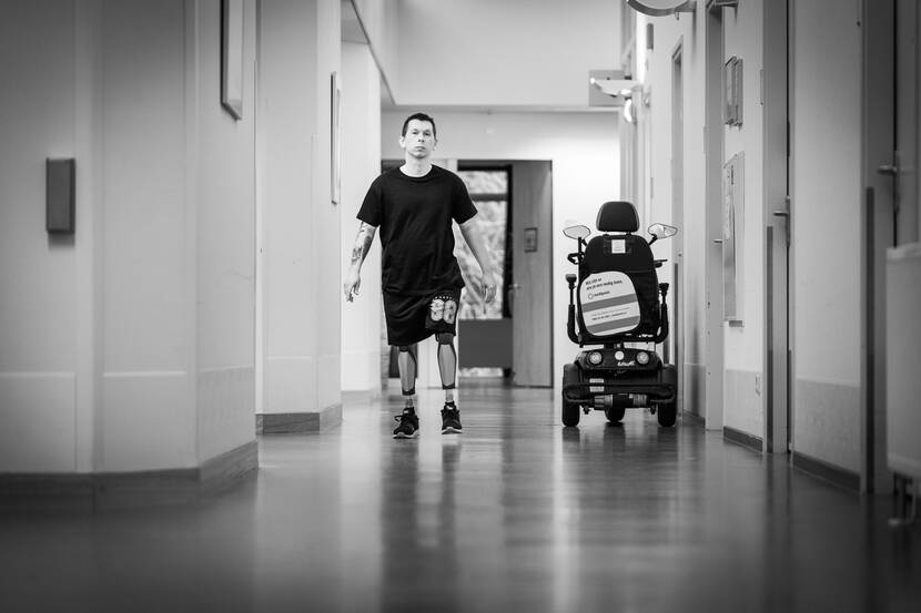 Alttekst: Illia loopt op zijn kunstbenen door een gang, rechts staat zijn scootmobiel.