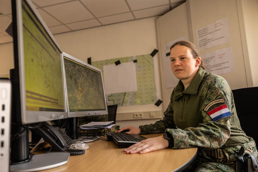 Militair in tenue zit aan een bureau achter 2 computerschermen.