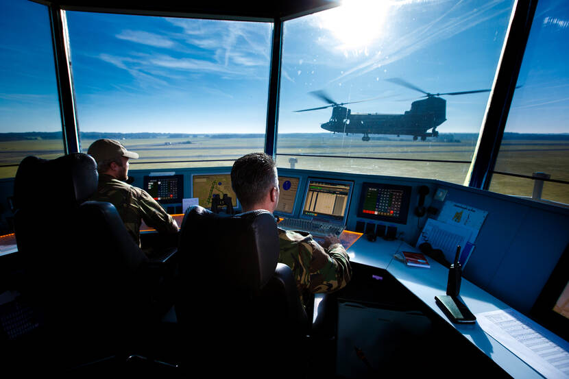 Twee luchtverkeersleiders aan het werk in een luchtverkeerstoren, buiten vliegt een Chinook voorbij.