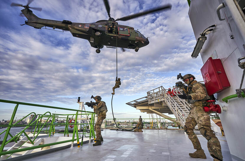 Special Operations Forces-militairen staan met getrokken wapen op het dek van een schip; op de achtergrond daalt een militair via een touw af uit een Cougar.