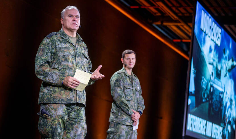Twee mannen in militair tenue op een podium naast een groot scherm.